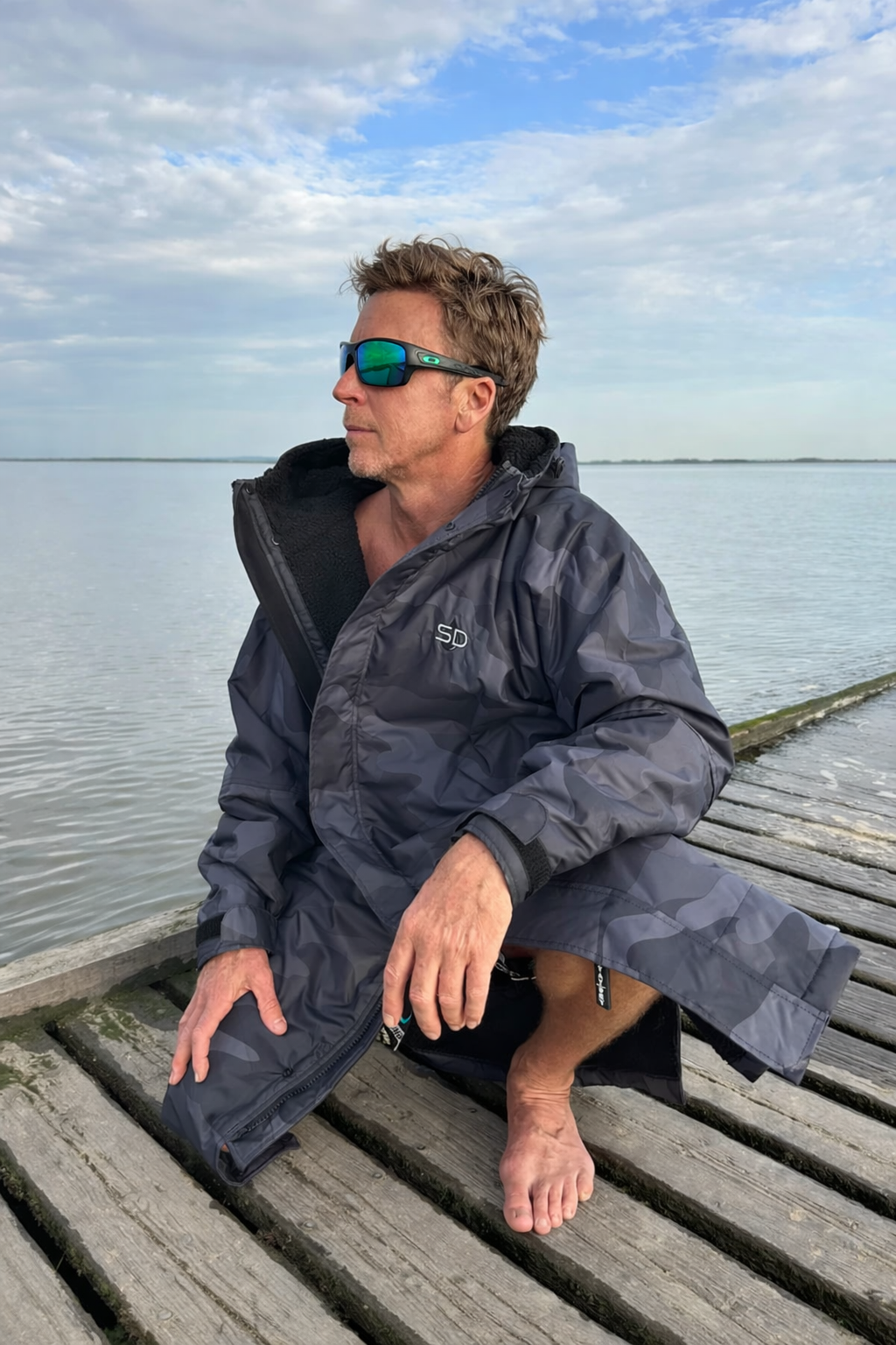 Person wearing a changing robe sitting on a wooden dock by a body of water.