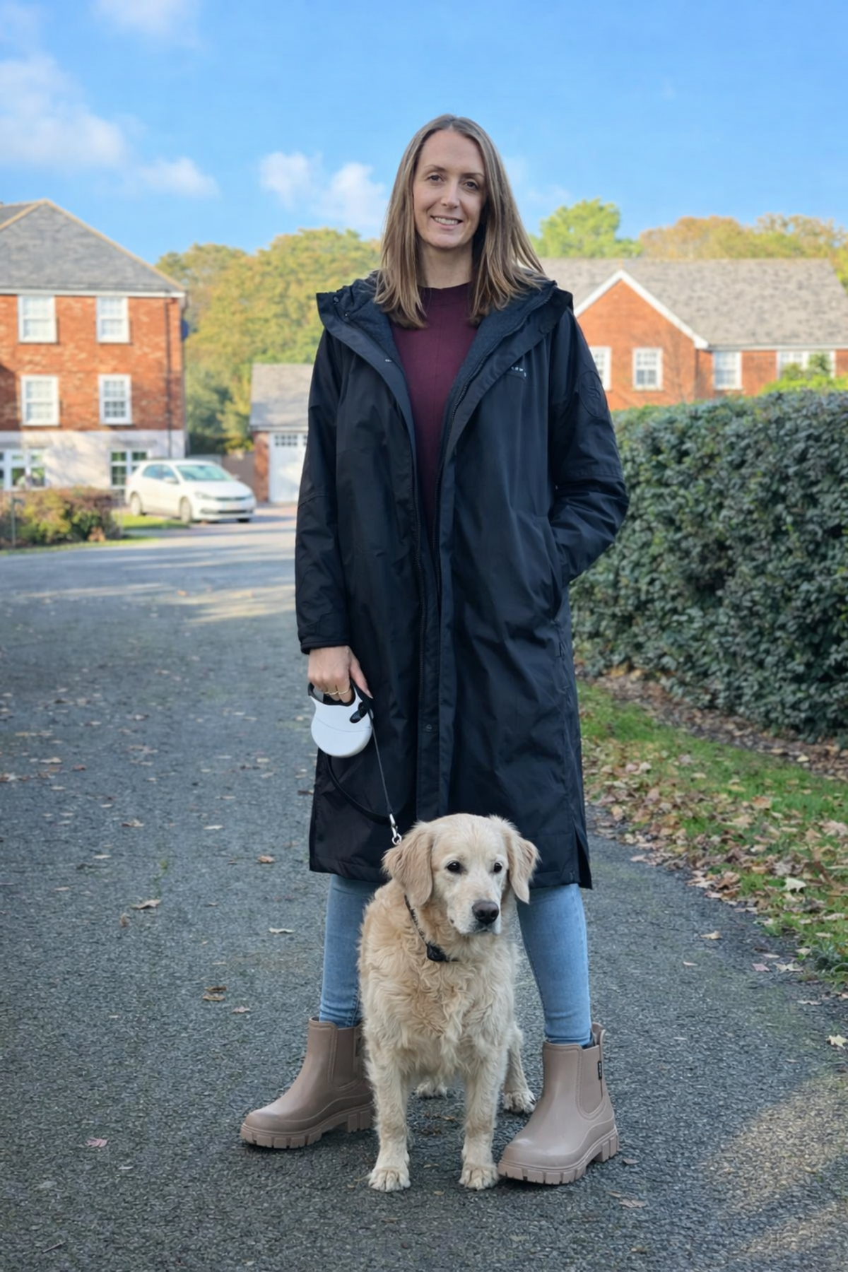 Woman in a long dark coat standing with a dog on a residential street.