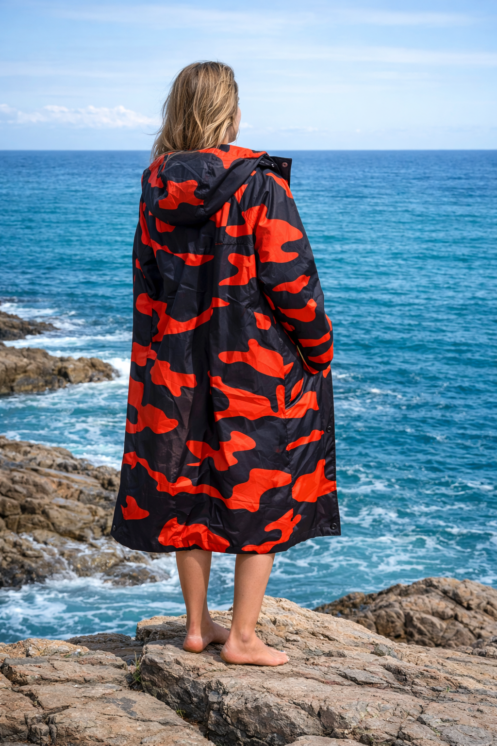 Person wearing an orange and black camouflage hooded blanket standing on rocks by the ocean.