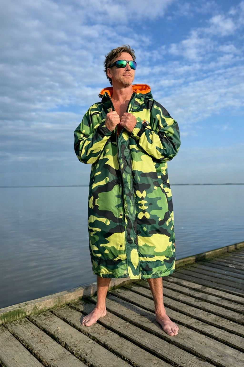 Man wearing a green camouflage hooded towel on a wooden dock with a blue sky and water background.