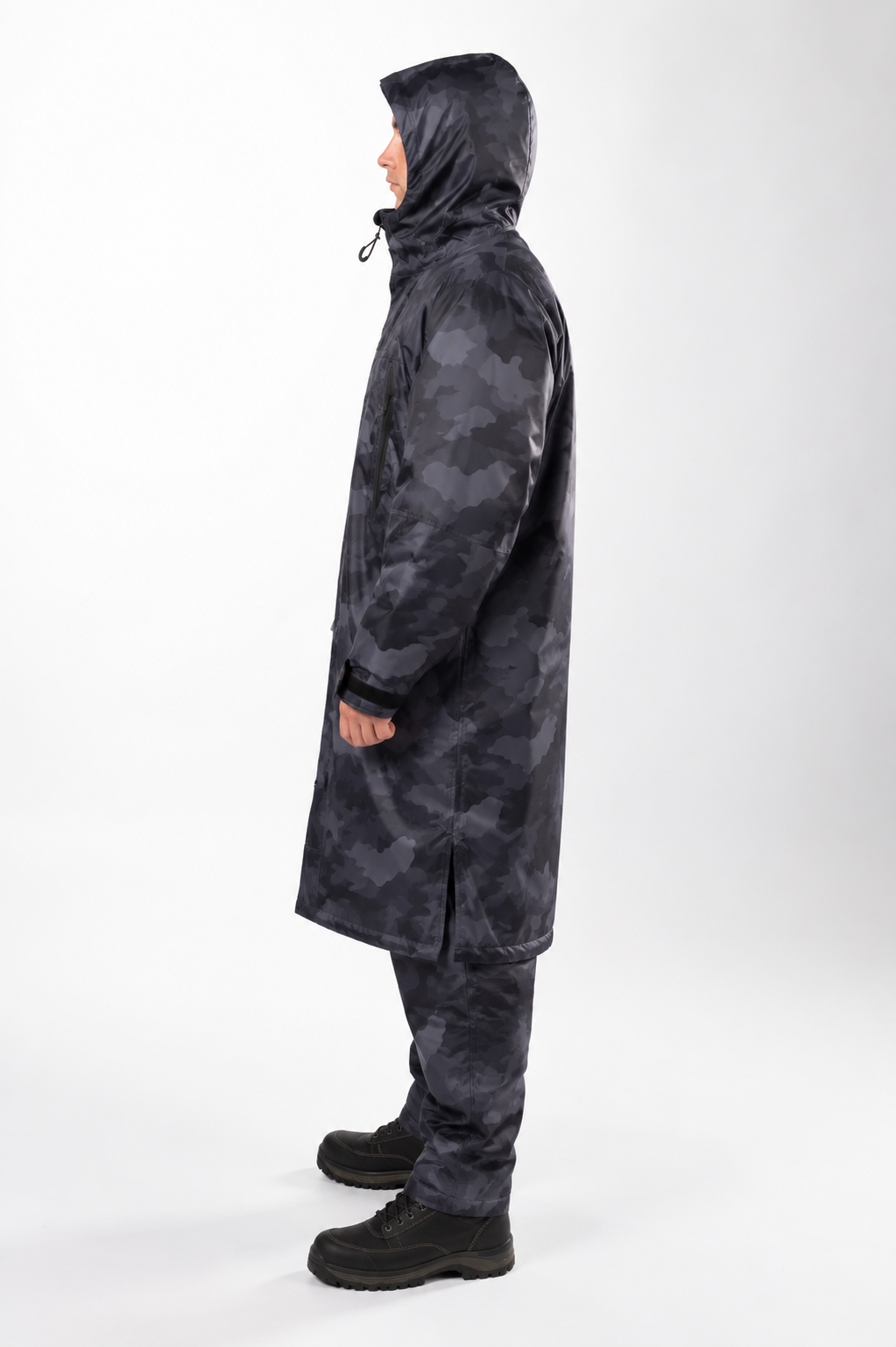 A person shown in profile wearing a long hooded fishing stay dry coat and matching pants in a dark gray and black camouflage pattern, standing against a light gray studio background.