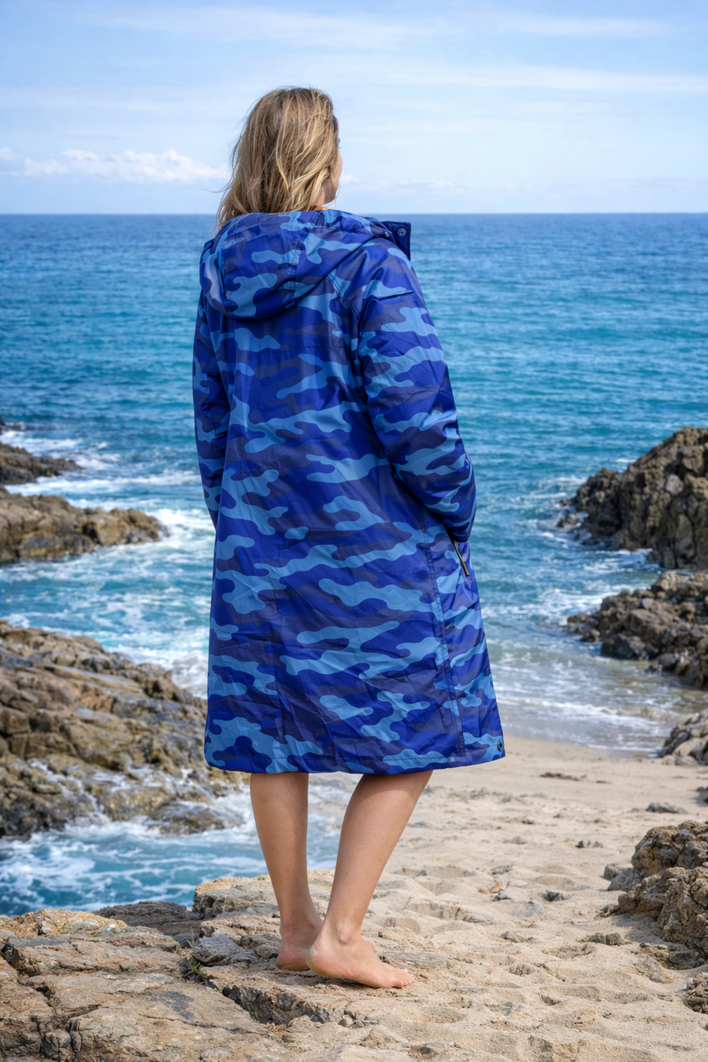 Person wearing a blue camouflage robe standing on a rocky beach with ocean waves in the background
