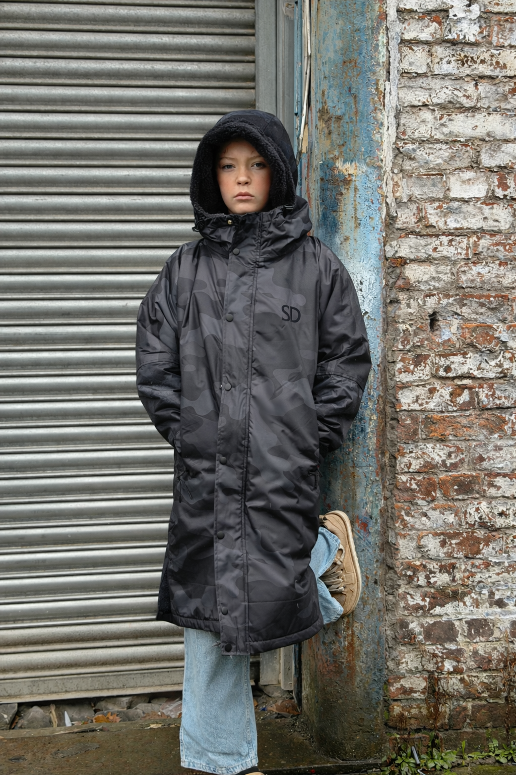 Person wearing a black raincoat standing against a textured wall and metal shutter.